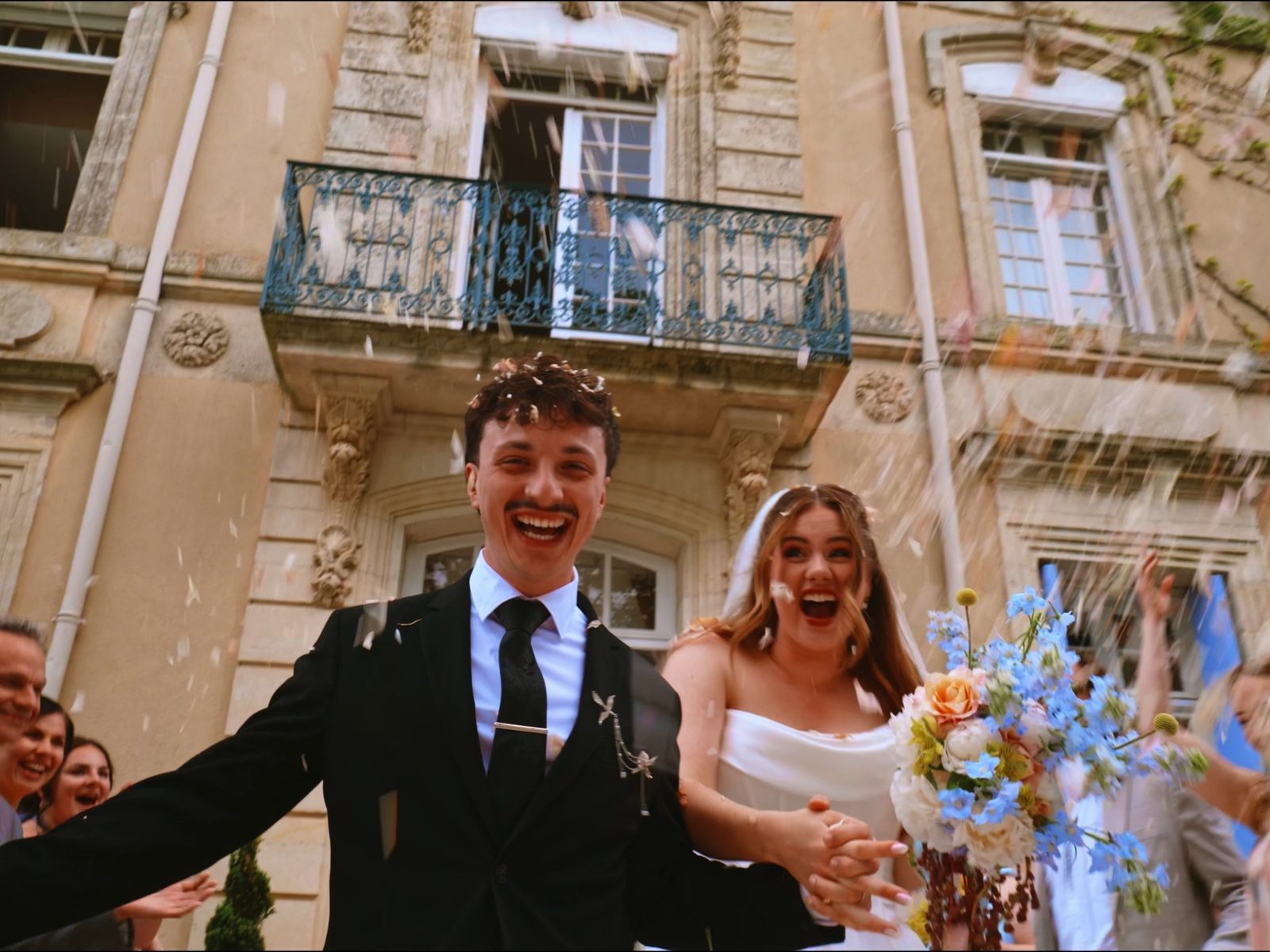 Newlyweds laughing as confetti falls in front of a stone château with a blue balcony