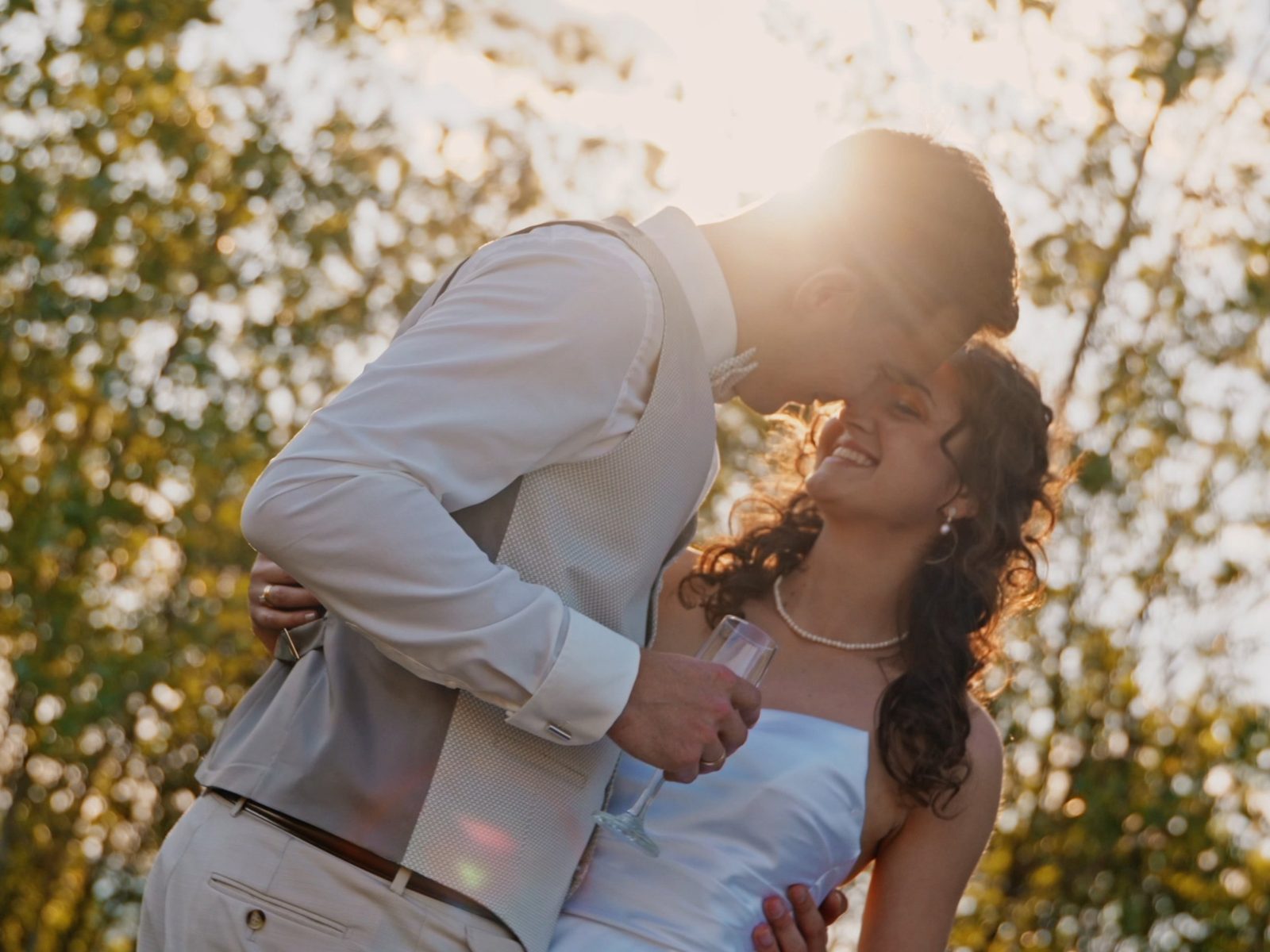 Groom leaning in to kiss his bride, backlit by golden sun through the trees, champagne glass in hand