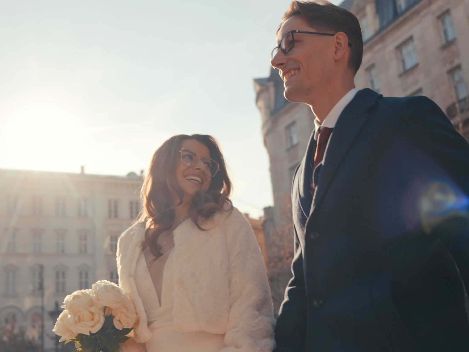 Smiling bride in a white fur wrap holding roses, looking up at her groom in a Budapest street