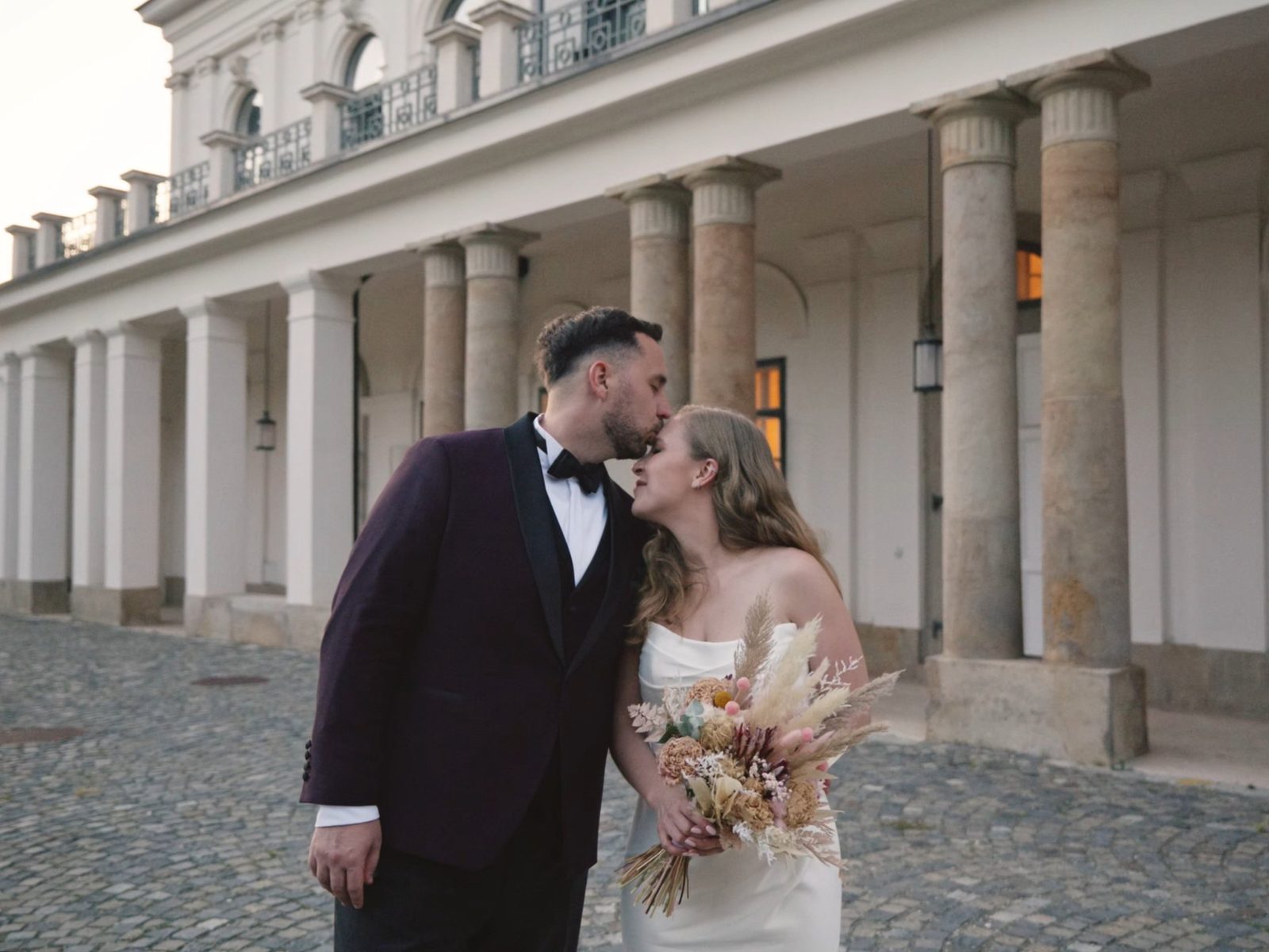 Groom kissing his bride on the forehead in front of a columned castle courtyard