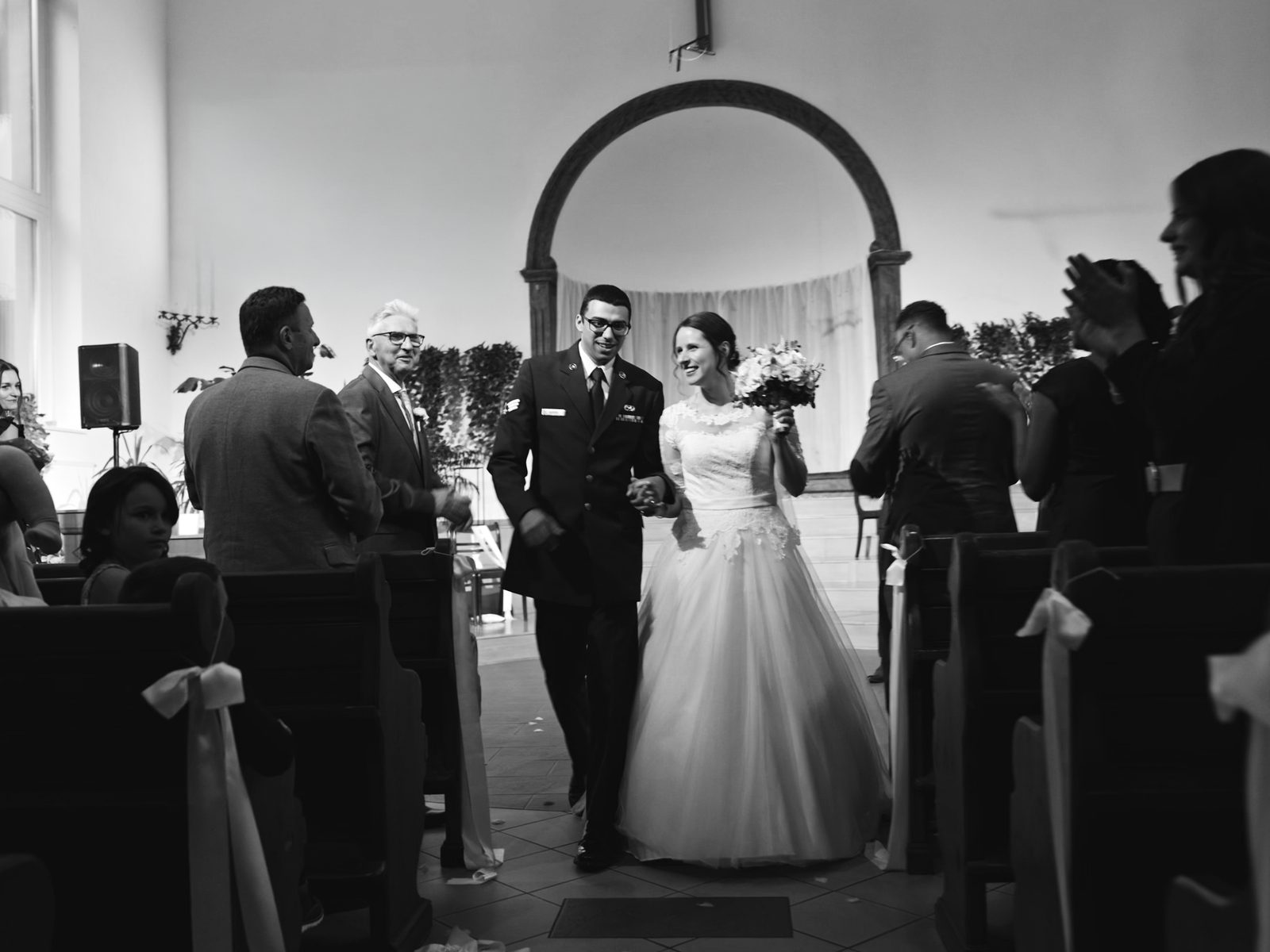 Black and white photo of a bride and groom walking up the aisle as guests applaud